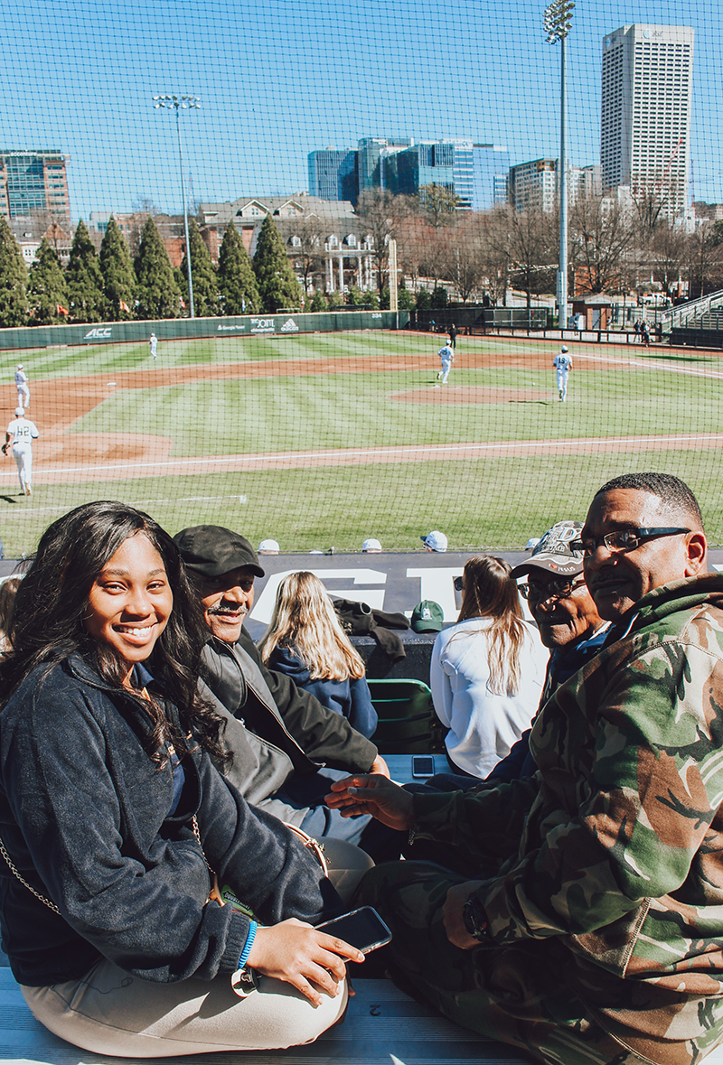 Smiling African American grandparents, father, and granddaughter with a baseball field behind them.