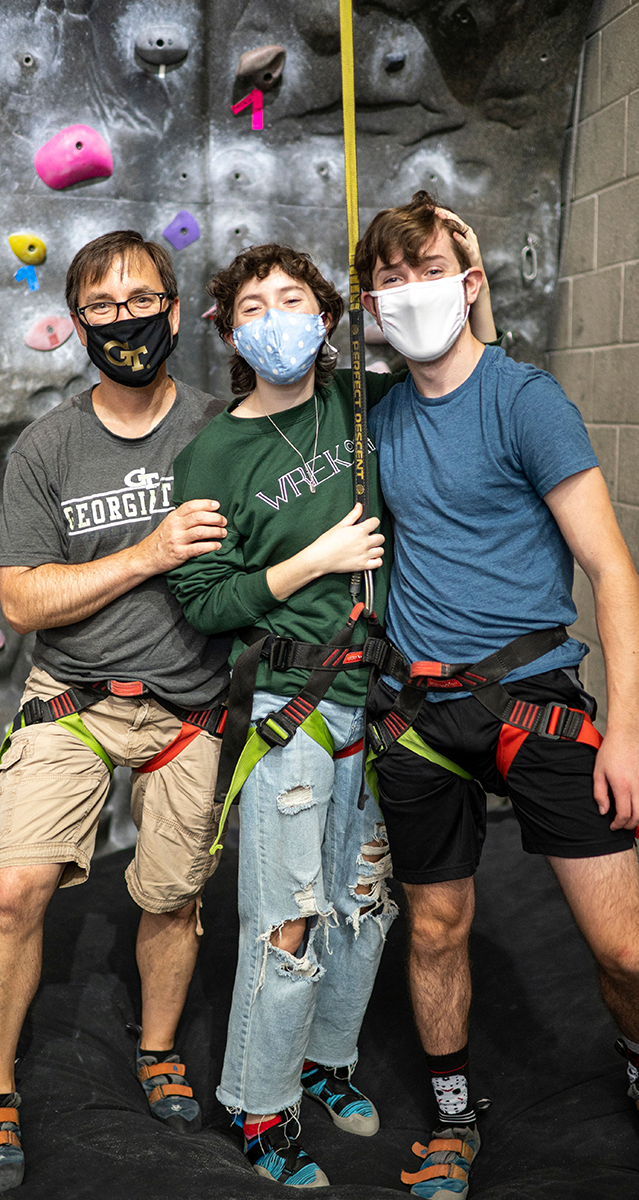 Father and 2 sons wearing masks and climbing gear in front of a rock wall.