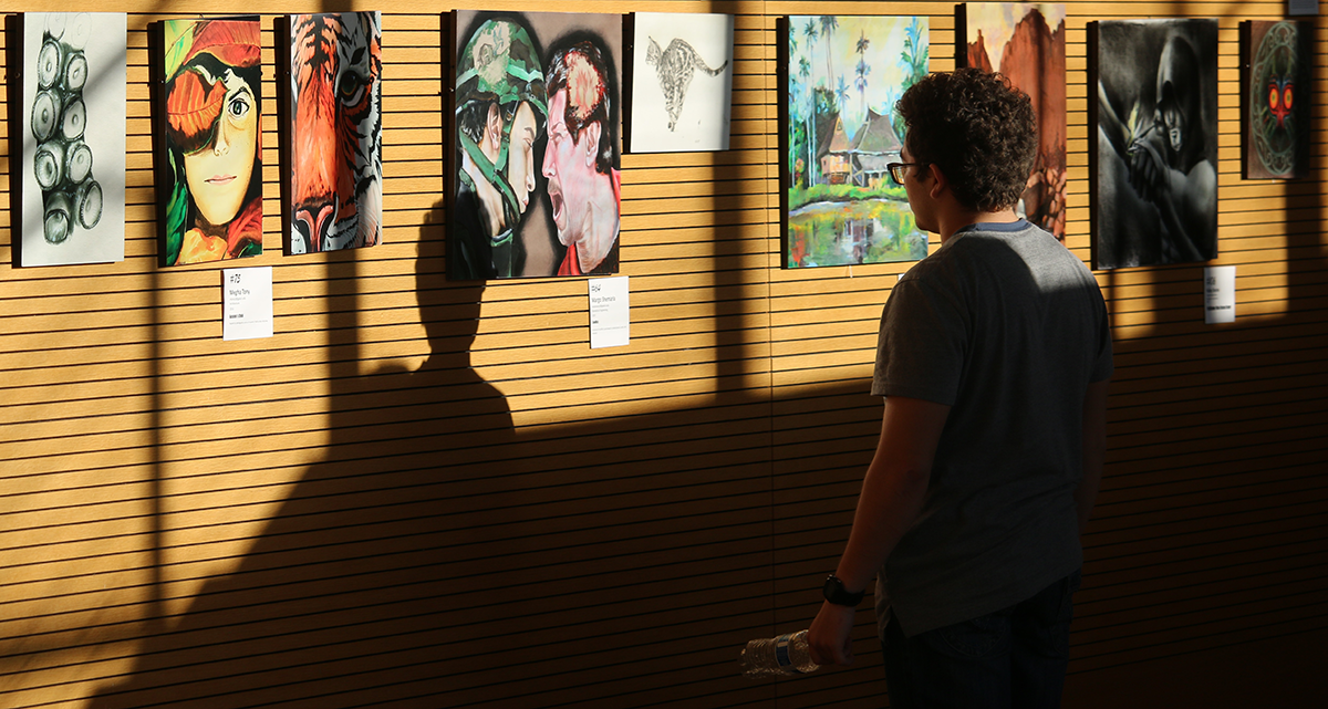 A wall full of paintings and a male student with his back to the camera looking at one.