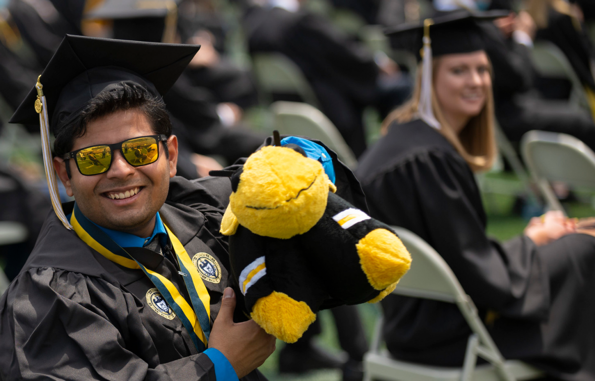 Asian male grad wearing cap and gown holding a soft plush Buzz.