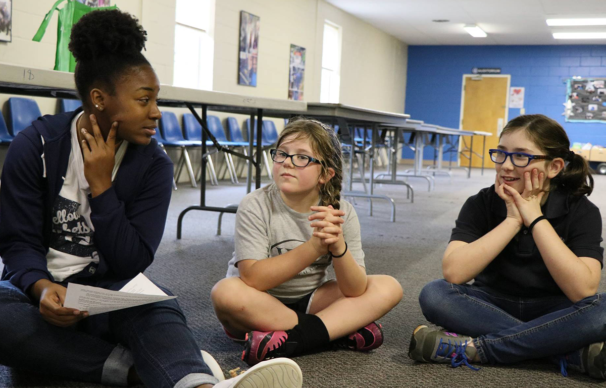 Black female college student sitting down on the floor with 2 white grade school girls.