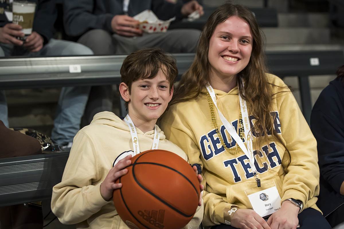 A young woman wearing a georgia tech sweatshirt sits next to her brother who is holding a basketball they are both smiling