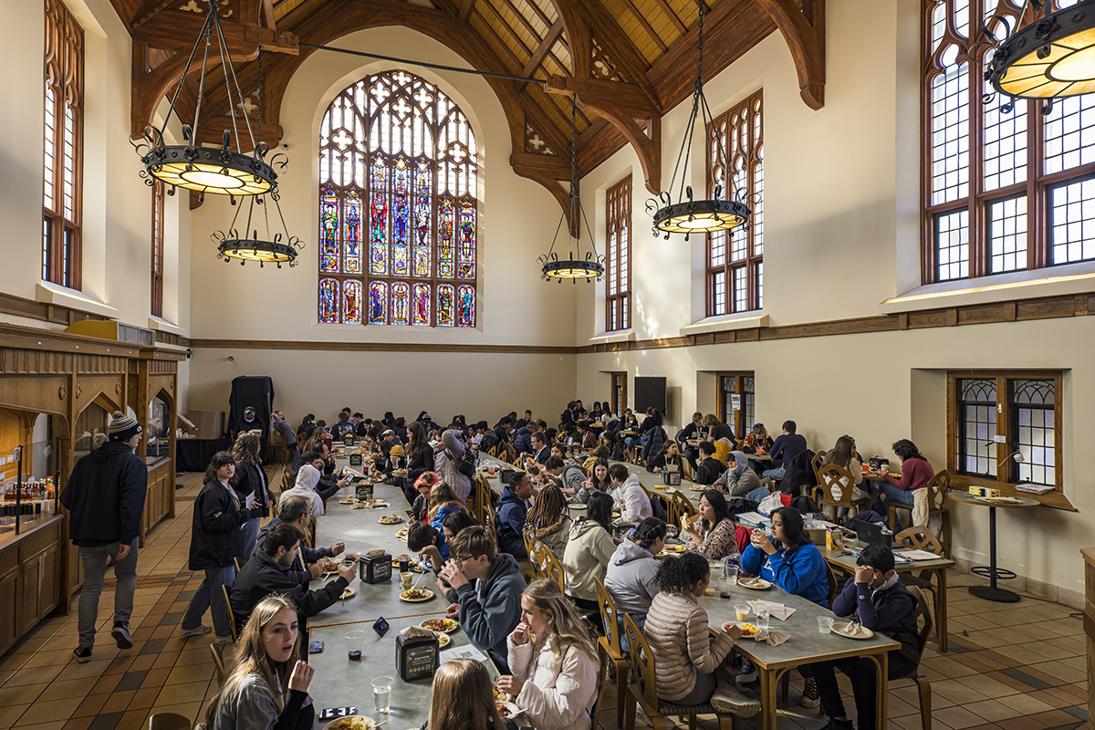 Hundreds of students sit and eat in a  cavernous dining hall with gothic arches and stained glass windows