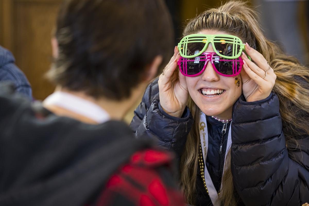 A young woman wears two pairs of flourescent sun glasses and is smiling at how silly she is