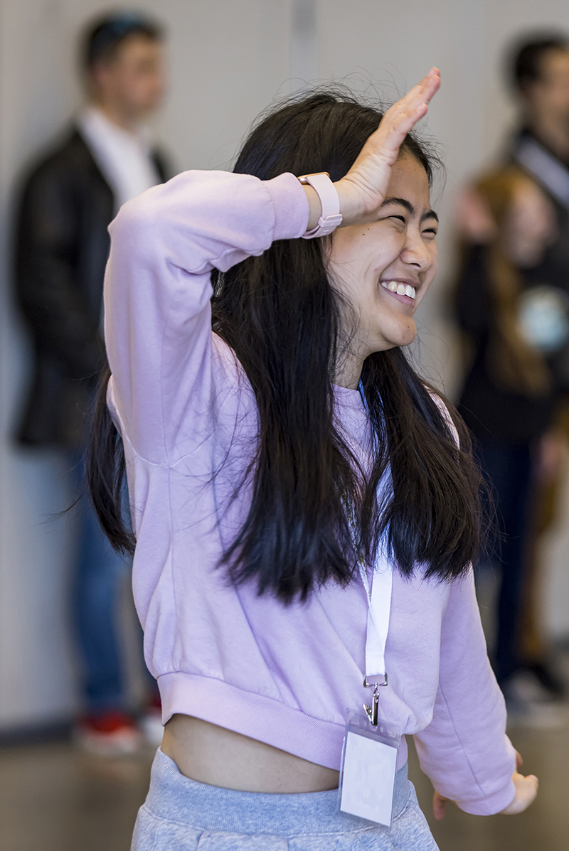 A young woman with long dark hair wearing a pink sweatshirt smiles as she does dance moves