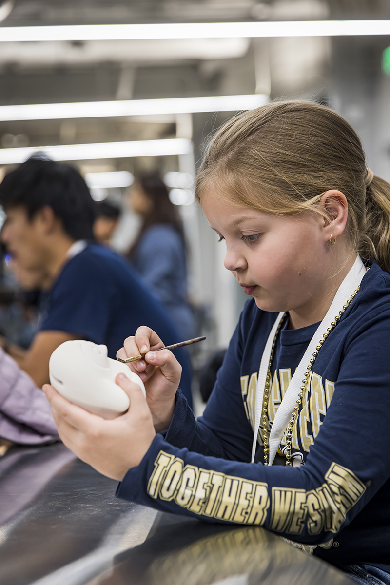 A young girl wearing a georgia tech shirt intently looks at the pottery in her hand as she paints it
