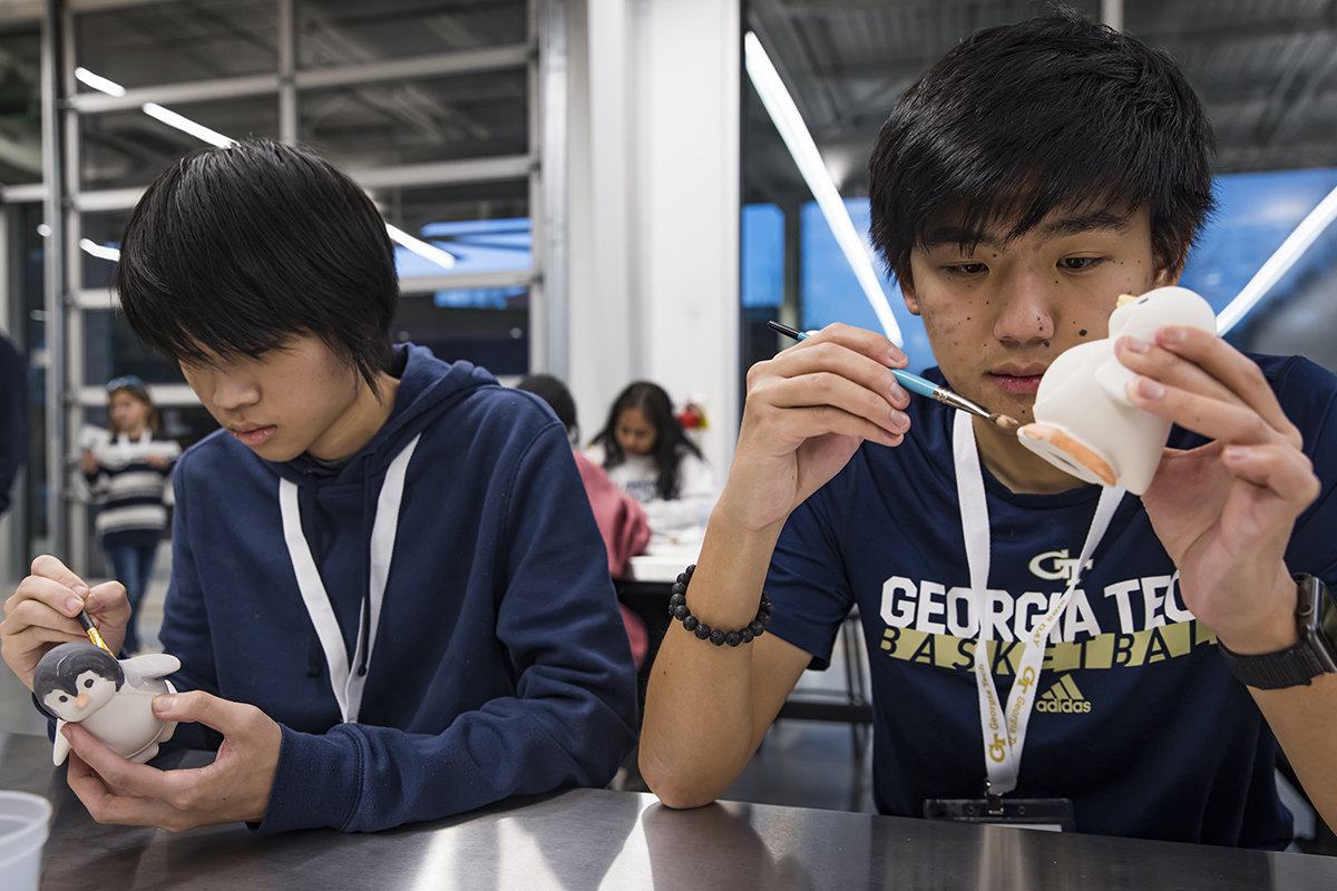 Two young men, each with dark hair and wearing blue sweatshirts, are concentrating on painting the pottery in their hands