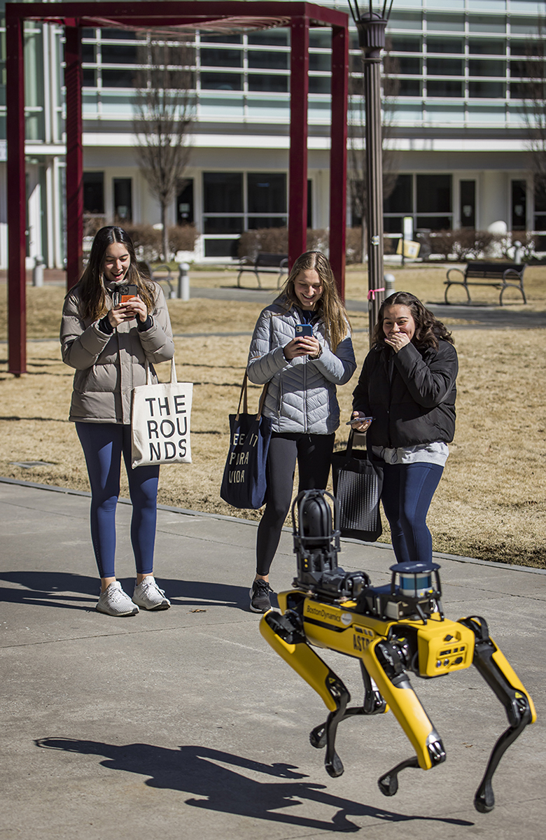 three young women laugh, smile, and take photos of a robotic dog walking on the pathway next to them