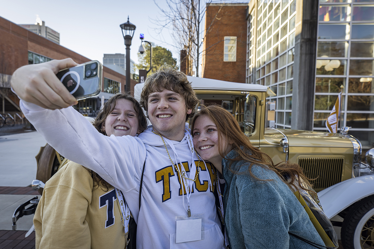 Three young people take a selfie in front of a gold and white Model A sport coupe