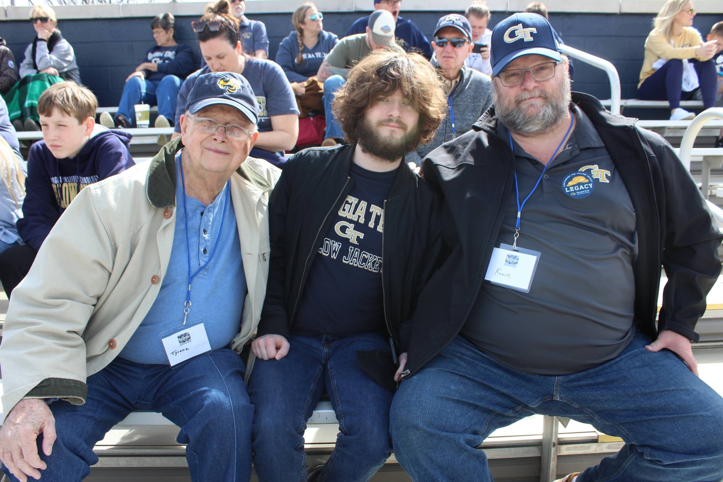 three generations of men sit in seats at a baseball stadium, all wearing Georgia Tech hats or shirts