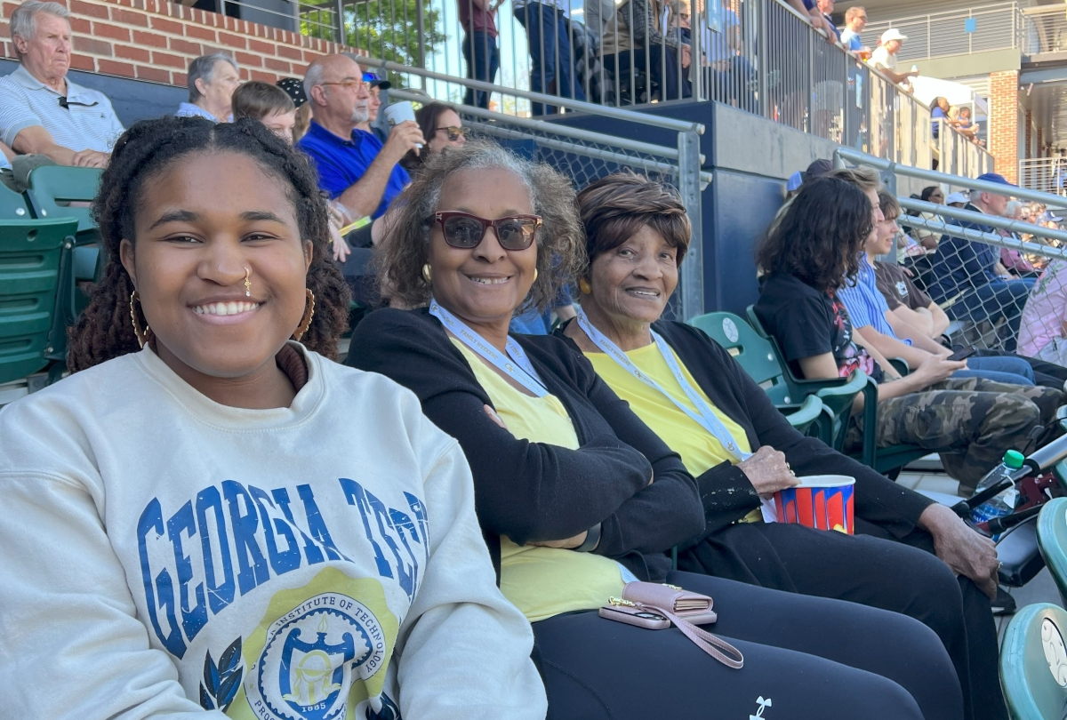 three generations of women, college student, mother, and grandmother, sit in an outdoor stadium at a baseball game