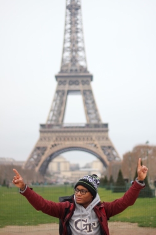 a photograph of a young man standing with his arms outstretched in front of the Eiffel Tower in Paris, France.