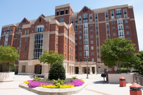A large brick building stands in the sunshine, fronted by tidy gardens and seating areas.