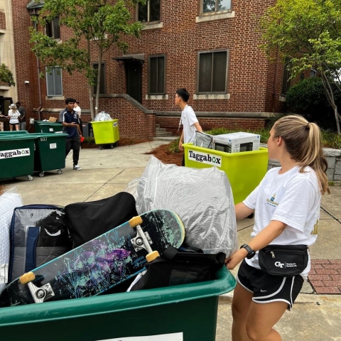 a young woman in white t shirts and navy shorts is pulling a large green bin filled with personal items as she moves out of her dorm